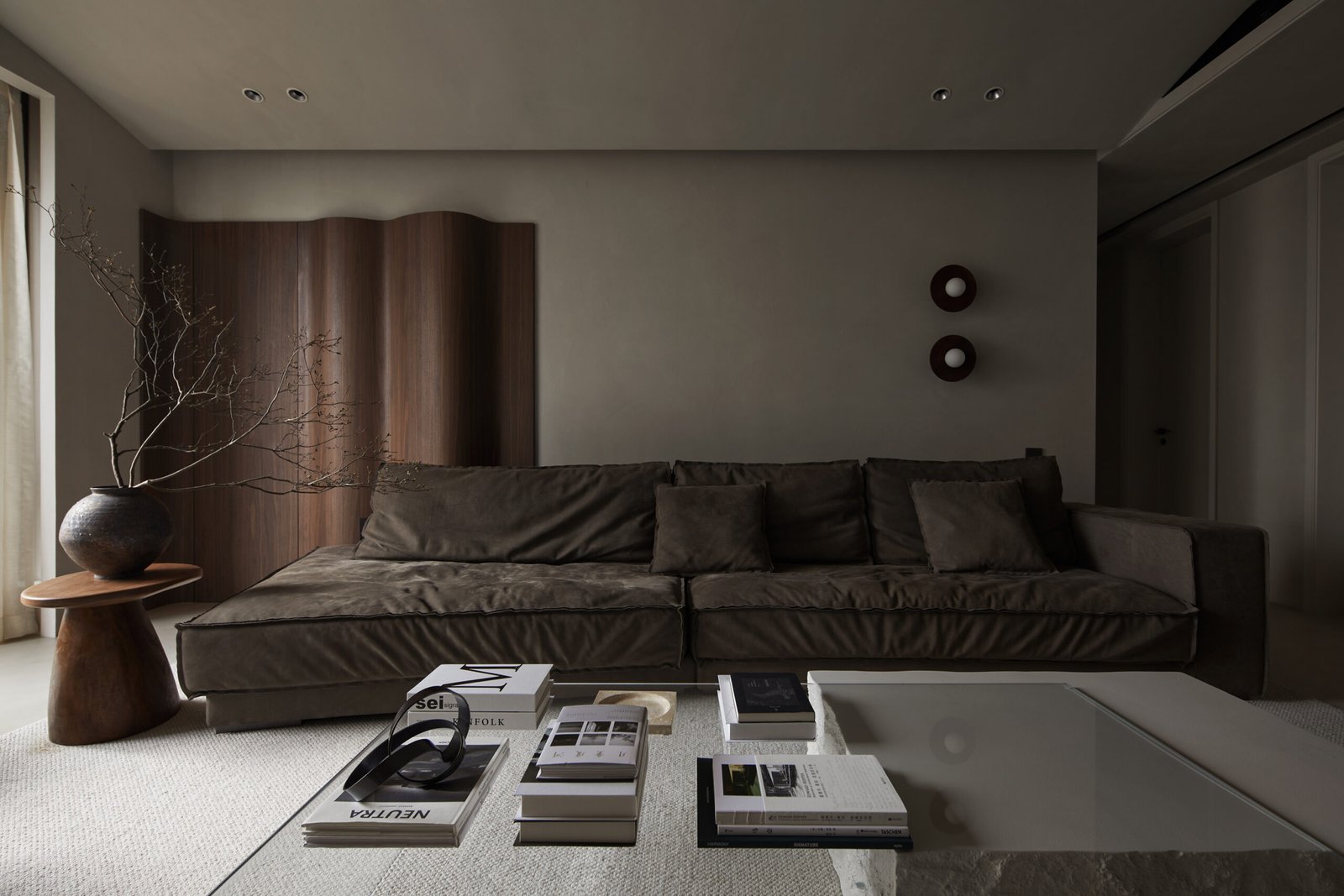 A modern minimalist living room featuring a low-profile dark brown velvet sofa, a wavy wood room divider, and a stone coffee table styled with art books.