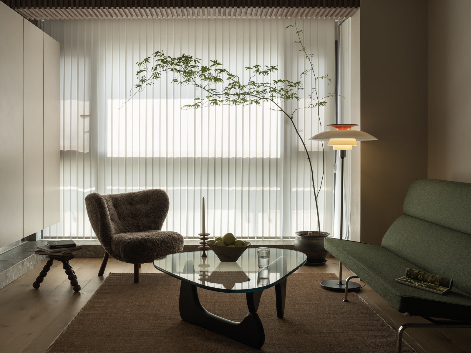 Compact living room with sheer vertical blinds, a sculptural glass coffee table and a slim potted maple